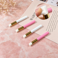 Four small makeup brushes with pink and white handles and gold ferrules arranged on a pink surface, with a circular inset showing a finger holding one brush.