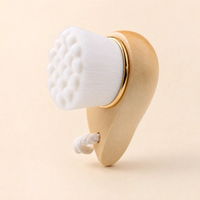 A wooden facial massage brush with a white bristle head, a gold metal accent ring, and a white hanging cord, shown on a solid beige background.