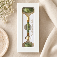 A jade green and gold facial massage roller presented inside a white gift box, styled on a beige linen fabric with small white flowers and a ceramic dish.